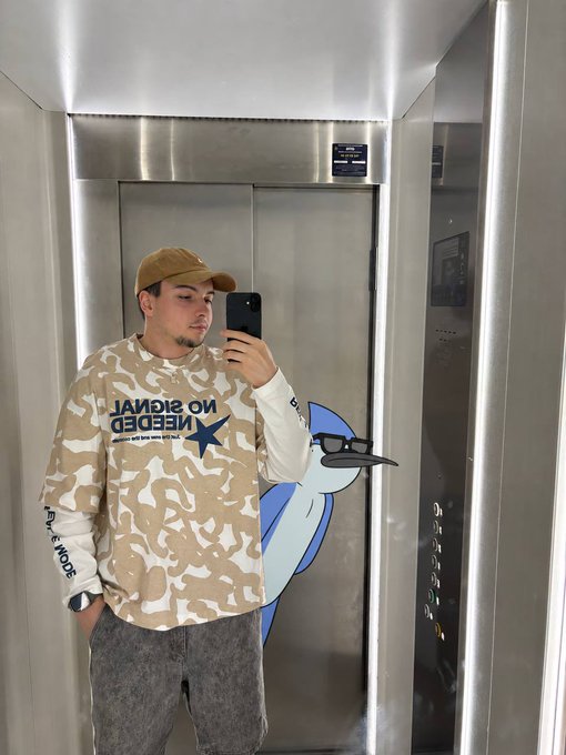 A man wearing a beige cap and a tan and white camo sweatshirt with "NO SIGNAL NEEDED" text takes a mirror selfie in an elevator while holding a smartphone.