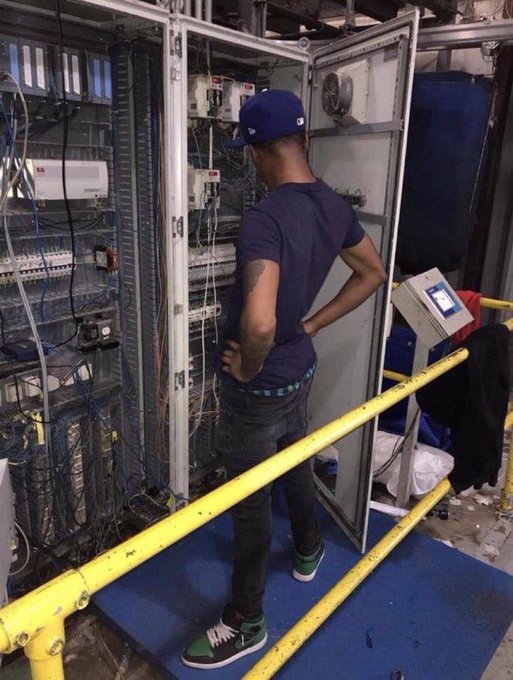 A young man in a blue cap and dark t-shirt working on or inspecting an open telecommunications/electrical server rack with extensive wiring, standing on a blue platform with yellow safety railings.