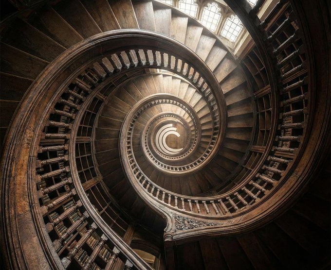 A downward view into an ornate wooden spiral staircase with intricate railings, illuminated by light streaming through windows at the top.