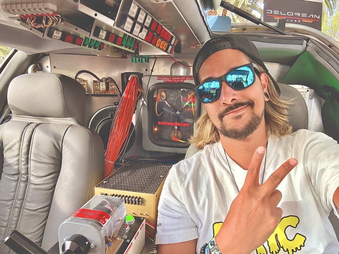 A man with sunglasses and a backwards cap flashes a peace sign from inside a heavily modified Delorean, where the signature gull-wing door and interior control panels reveal the futuristic-looking array of wires, switches, and circuit boards reminiscent of Doc Brown's time machine. The "Delorean Cars" emblem is visible on the rear, playfully tying into the "GM future" caption.