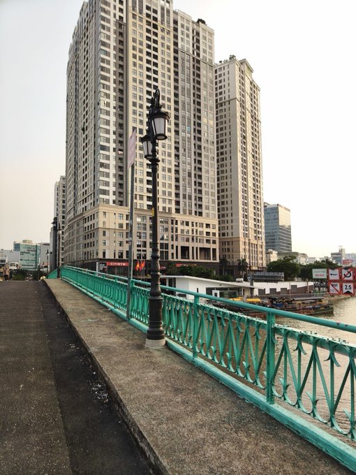 A walkway beside a river lined with a turquoise railing and an ornate lamppost, with a tall beige high-rise residential building dominating the background.