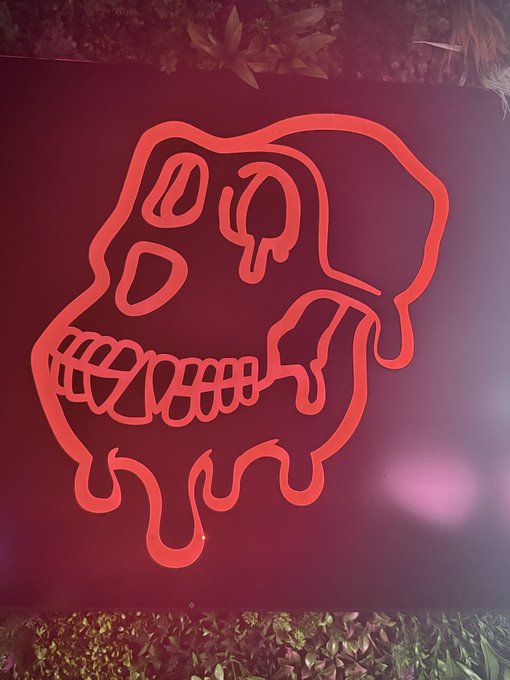 A red neon sign featuring a stylized skull with dripping, melting features is mounted on a wall, surrounded by green foliage.