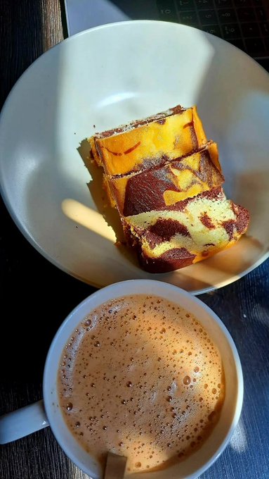A close-up photo of a marble cake slice on a white plate next to a cup of coffee, with sunlight casting shadows.