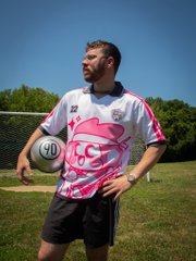 A man wearing a white, pink, and purple sports jersey with '22' and 'CCSC' stands on a grass field holding a soccer ball.