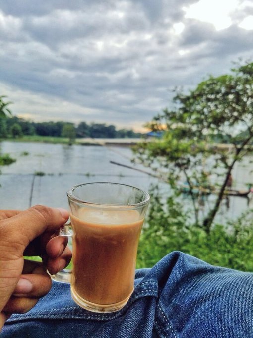 The image shows a person holding a glass of coffee or tea with milk and sugar, sitting outdoors near water. The scene appears to be set by a lake or river, creating a serene atmosphere for enjoying a hot beverage. The photo also includes text that reads "Good morning Don't just exist - grow, learn and shine." This message is related to the person's coffee or tea drink in the image, suggesting they are taking time to enjoy their drink while appreciating nature and personal growth.