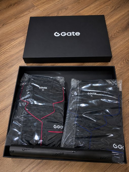 A product photo of an open GGate branded box on a wooden floor, containing black clothing items in plastic packaging and a rolled item, marked with M and XL sizes.