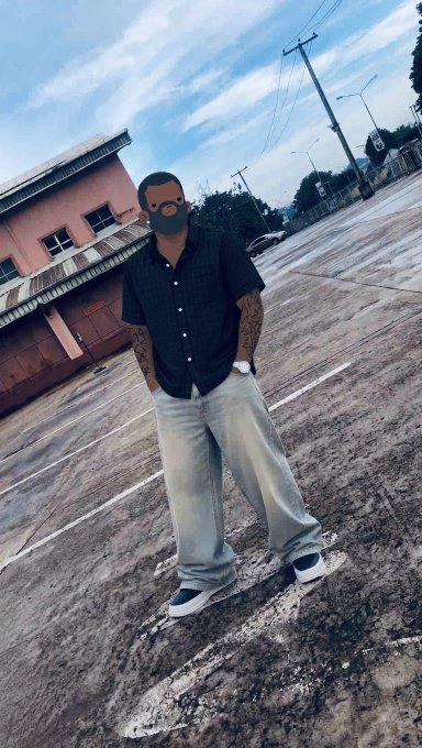 A man wearing a black face mask, a dark short-sleeved button-up shirt, and baggy light blue jeans stands on a road near a pink building, with his face covered by a gray icon.
