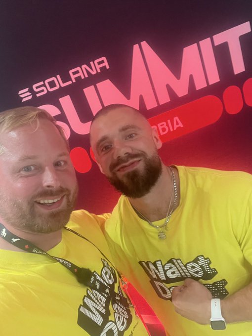 Two men in matching yellow Waldom DAO shirts smile for a selfie at the 'SOLANA SUMMIT SERBIA' event.