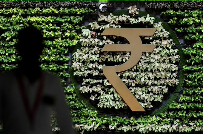 The Indian Rupee symbol (₹) stands out in gold against a lush wall of green and white plants, with a silhouetted figure in the foreground wearing a lanyard. The image relates to India's growing fintech and tokenisation efforts, visually symbolizing the Indian currency central to these new digital asset developments.