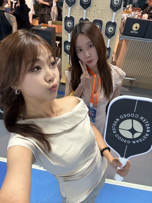 Two young women pose playfully for a selfie at what appears to be an Aster DEX Glow Pickleball event, with one holding a black paddle branded "#R3T4 #CODED #W3LC0M3D" while the other flashes a peace sign in a white lace dress.