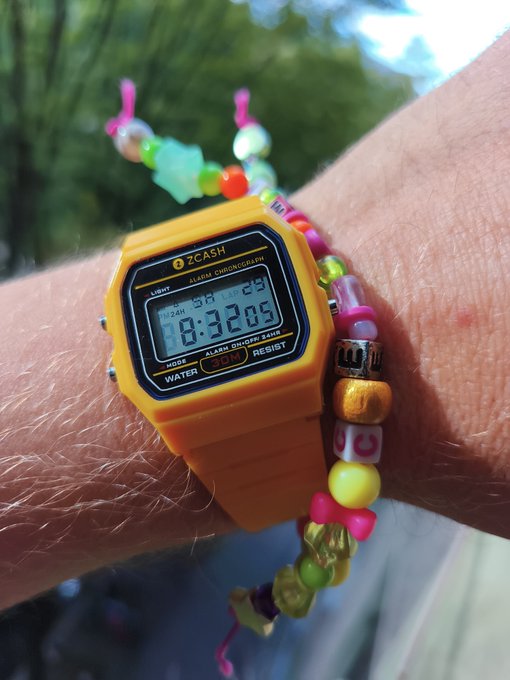 An orange ZCASH digital watch on a wrist displays the time 10:32 with date "29" and is paired with a colorful beaded bracelet.