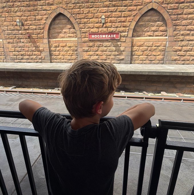 A young boy wearing a dark gray shirt leans on a black metal railing, looking toward a 'HOGSMEADE' station sign mounted on a brick wall.