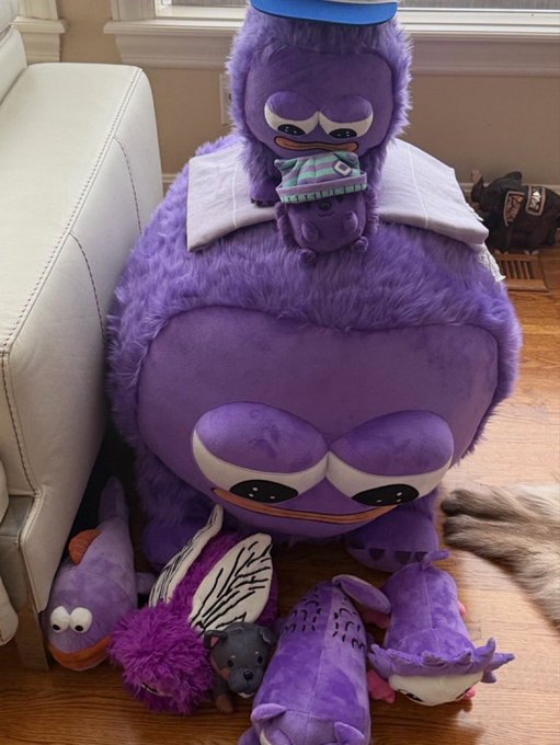 A large purple plush toy with a grumpy expression and a blue cap sits on a wooden floor, surrounded by several smaller plush creatures.