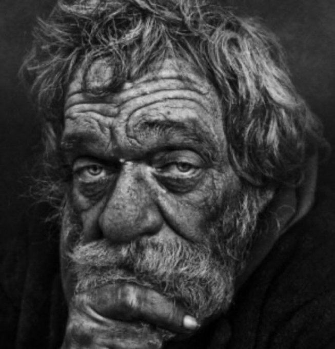 A black and white portrait photograph of an elderly man with messy hair and a weathered face, resting his hand on his chin in a contemplative pose.