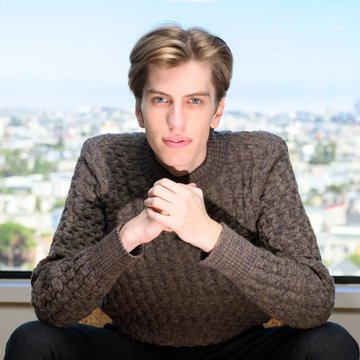 A man with light brown hair and blue eyes sits wearing a textured brown knit sweater, with a cityscape visible through a window in the background.