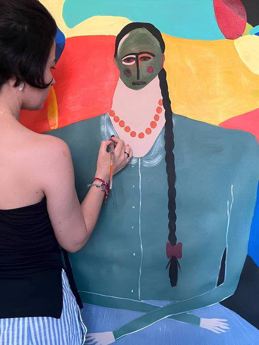 A woman with dark hair in a strapless top paints a colorful, stylized portrait of a figure with a green mask, orange beaded necklace, and long dark braid.