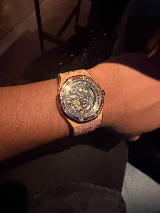A first-person photo of a person's wrist wearing a luxury rose gold skeleton-style automatic watch with a visible tourbillon mechanism and a bezel set with rainbow-colored gemstones.