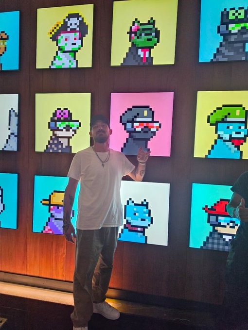 A man wearing a white t-shirt and cap stands in front of a wall displaying colorful pixel-art portraits of characters including Princess Peach, Shrek, and Astro Boy.