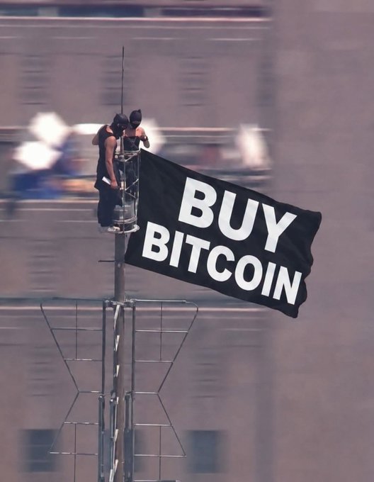 Two people climbing on top of a tall antenna/broadcast tower, unfurling a large black banner reading 'BUY BITCOIN' against a city backdrop.
