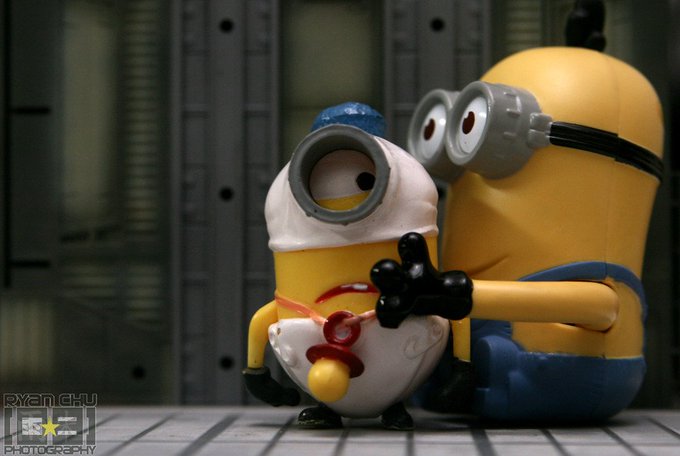 The image features two toy Minions, one wearing a white hat with a red nose and the other wearing a yellow hat with black eyes, sitting next to each other. The Minions are holding a pacifier between them, which adds an element of humor to the scene. The background is blurred, drawing attention to the Minions and their playful interaction.