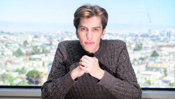 A man with brown hair wearing a dark gray sweater sits with his hands clasped together, appearing to be in an interview setting with a cityscape visible through the window behind him.