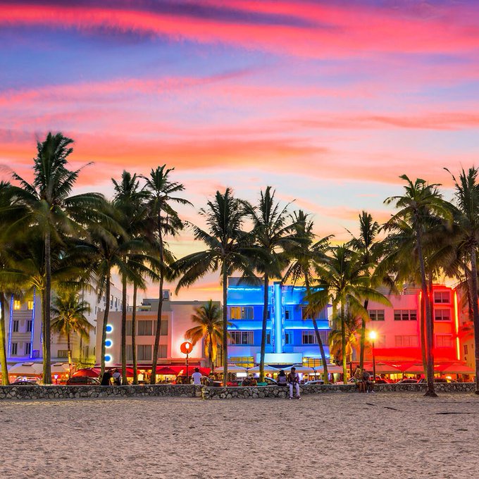 A vibrant sunset photo of Ocean Drive in Miami Beach, featuring palm trees silhouetted against pink and purple skies and colorful Art Deco buildings lit with neon.