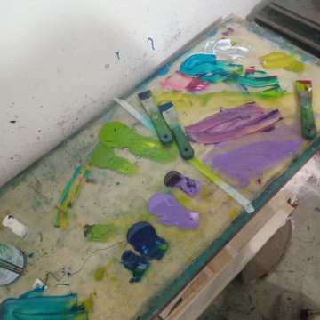 A messy artist's palette covered in smears of green, purple, and blue paint with several palette knives and brushes resting on it.