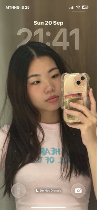 An iPhone lock screen selfie showing 'MTNNG IS 25' with the time 21:41 on Sun 20 Sep, featuring a young woman in a pink t-shirt taking the photo in a mirror.