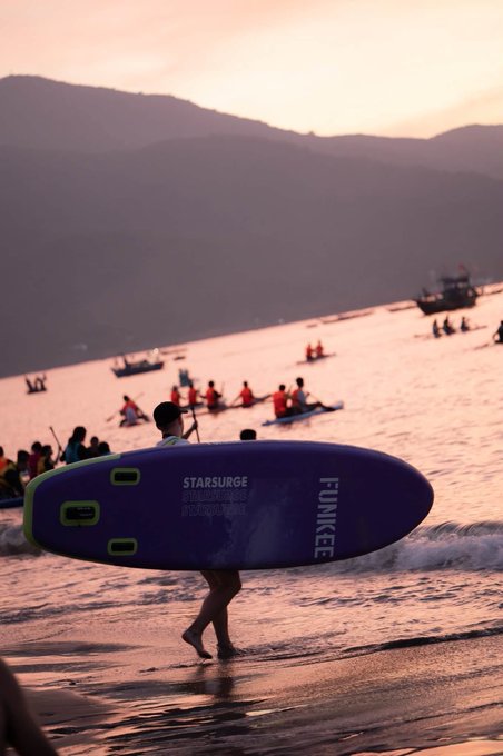 A person walks along a beach at sunset carrying a blue and green "FUNKEE" branded surfboard labeled "STARSURGE," with kayakers and boats visible in the water behind them.