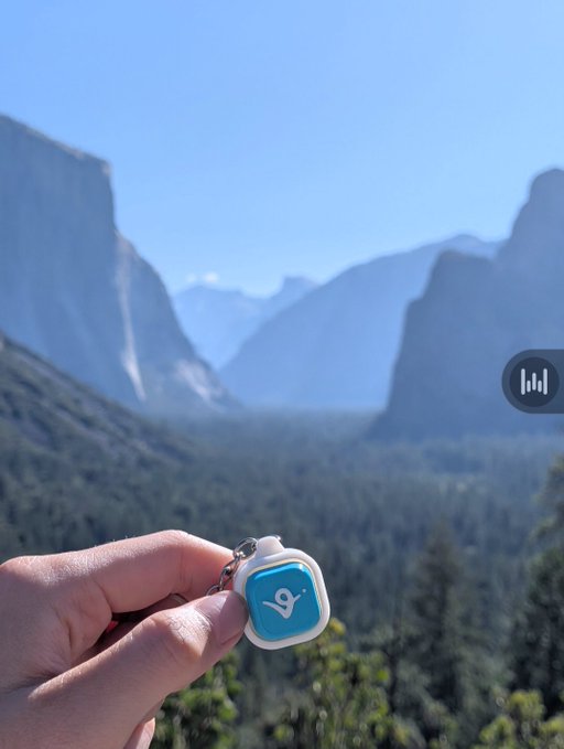 A hand holds up a small blue and white keychain with a location pin logo against a scenic backdrop of Yosemite Valley's granite cliffs and forest.
