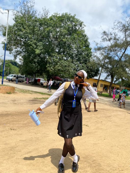 A photograph of a smiling young woman in a school uniform (white shirt, black vest, black skirt with cross patterns) and a white head covering, holding a light blue water bottle outdoors near trees.