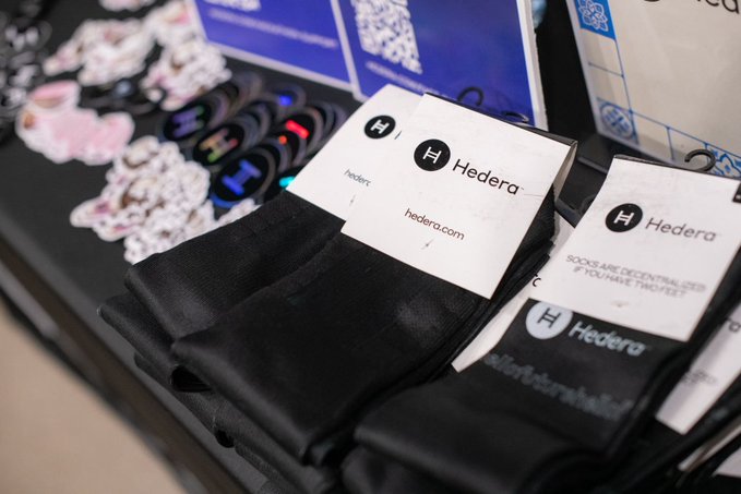 A display table features black socks with white tags reading 'Hedera' and 'hedera.com', alongside stickers and promotional materials.