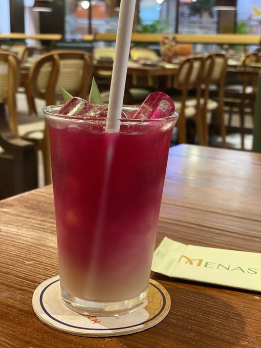 A vibrant pink and white layered iced drink with a straw sits on a coaster at a table, with a napkin displaying 'MENAS' partially visible.