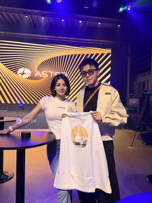 A woman and a man pose together holding a white t-shirt featuring the Aster logo, with the Aster DEX banner displayed on the screen behind them. The man thanks "CC" in the caption, suggesting she is the woman pictured, while promoting Aster's Korea-exclusive trading competition.