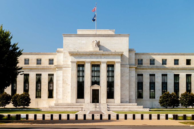 Exterior view of a large white neoclassical government building, likely the Federal Reserve, featuring tall columns, an American flag, and a central entrance.