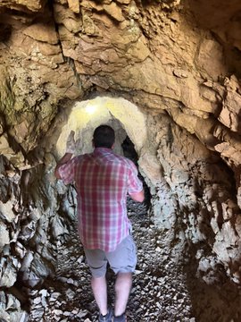 A person wearing a red and white plaid shirt and gray shorts stands facing a rock wall in a cave or tunnel.