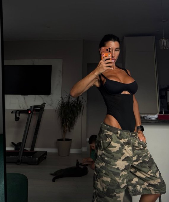 A woman takes a mirror selfie in her black cutout bodysuit and camouflage cargo shorts, with a treadmill and pet visible in the background of her modern home. The morning-themed image aligns with her "Good morning everyone" greeting, though it has no direct connection to the cryptocurrency hashtag.
