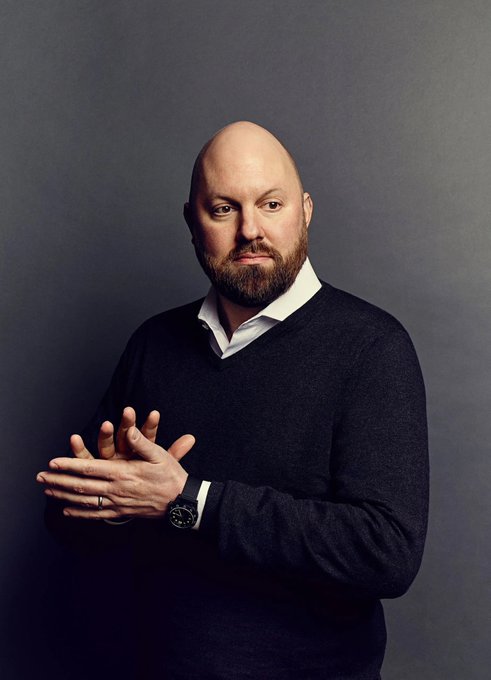 A studio portrait of a bald man with a beard, wearing a black v-neck sweater over a white collared shirt, posing with his hands clasped and wearing a wristwatch and ring.