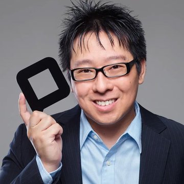 A smiling man with glasses and a light blue shirt holds up a black square object with a hollow center, wearing a dark blazer.
