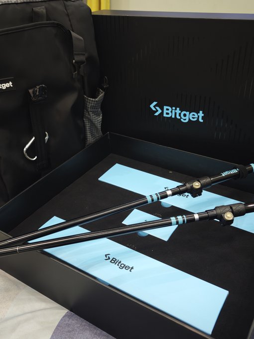A Bitget-branded backpack and a pair of trekking poles are displayed in their presentation boxes, featuring the "Bitget" logo and the company's signature blue arrow design, received as 8th anniversary swag gifts.