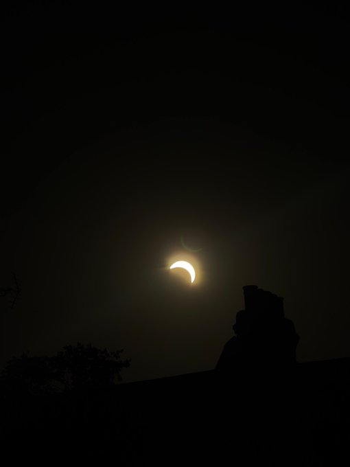 A crescent solar eclipse glows in a dark night sky above the silhouetted rooftops and trees below.
