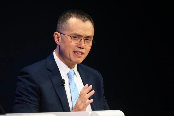 A photograph of Changpeng Zhao (CZ), the former CEO of Binance, wearing glasses, a dark suit, and a light blue tie, speaking at a podium.