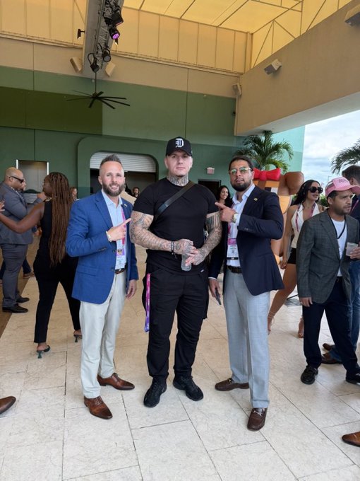 Three men pose together at what appears to be a formal event, with the central figure wearing a black cap, black t-shirt, and black pants, flanked by two men in blue blazers pointing at him.