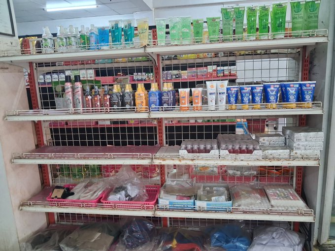Shelves in a convenience or general store stocked with various personal care products, toiletries, and bottled drinks.
