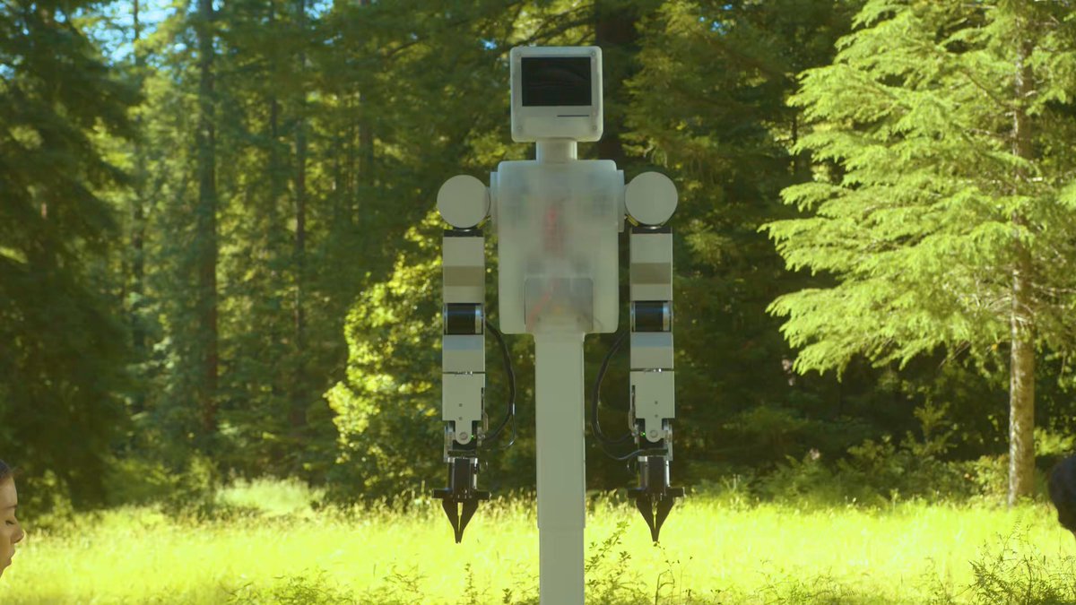 The image features a robot standing upright on its hind legs, situated outdoors in a park with trees surrounding it. The robot appears to be functioning as an information booth or display for an event.