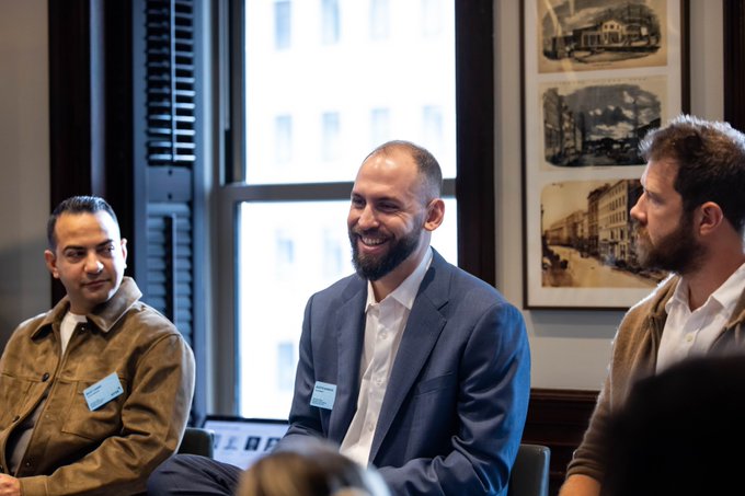 Three men seated at what looks like a panel discussion; the man in the center is smiling and wearing a blue blazer with white shirt and a name tag.