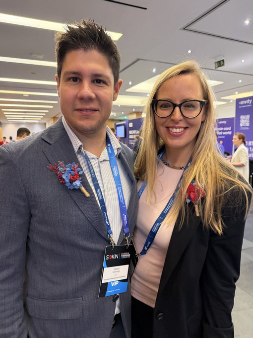 A photo of a man and a smiling woman at an event, both wearing "S-KIN" lanyards, with the man wearing a "VIP" badge and red floral boutonnieres.