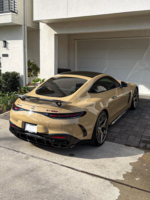 A tan/desert-colored Mercedes-AMG GT 63S sports coupe parked in a driveway in front of a modern house garage.
