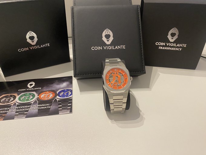 A Coin Vigilante "Transparency" branded watch with an orange dial and stainless steel bracelet rests on a display stand, accompanied by "COIN VIGILANTE" branded boxes and an instruction manual showing additional watch designs, matching the prize mentioned in the post.
