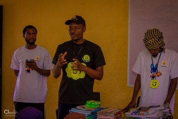 Three young men stand together at what appears to be a BNB-themed event, with one in the center wearing a black shirt and cap featuring a yellow smiley face logo while gesturing as if speaking, flanked by two others wearing white shirts and lanyards, all standing near a table displaying colorful items. The image relates to the post thanking @cz_binance and @GiggleMascotBNB by showing individuals associated with the BNB community, likely representing the Giggle Mascot BNB initiative.
